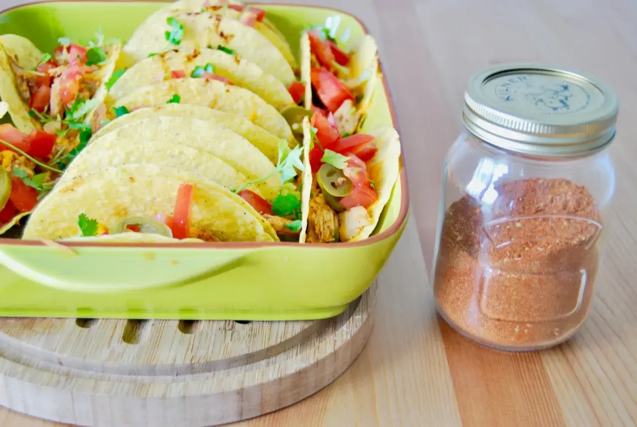 Domowa przyprawa do taco w słoiku obok talerza pełnego tacos z pomidorami i jalapeno. Lepsza niż gotowa z torebki!