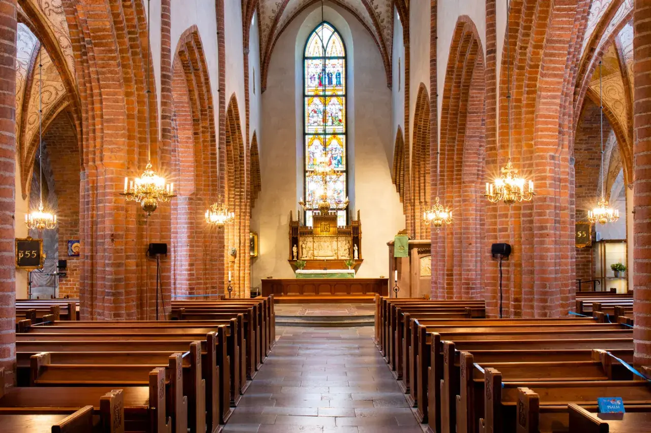 Interiör av Helga Trefaldighets kyrka med bänkar, tegelvalv och ett färgstarkt glasmålat fönster bakom altaret.