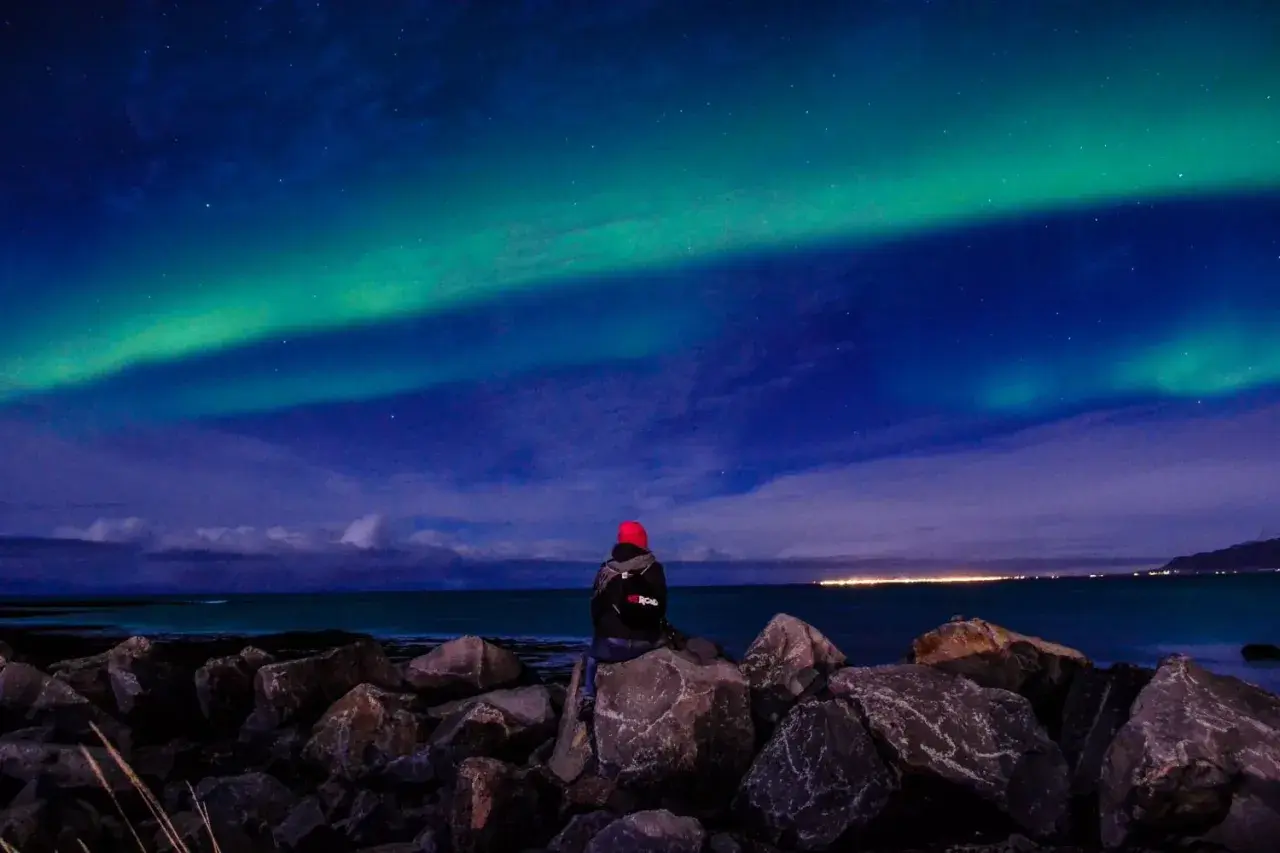 Person sitzt auf Felsen am Meer und blickt auf Nordlichter. Perfektes Wetter auf Island im September für dieses Naturschauspiel.