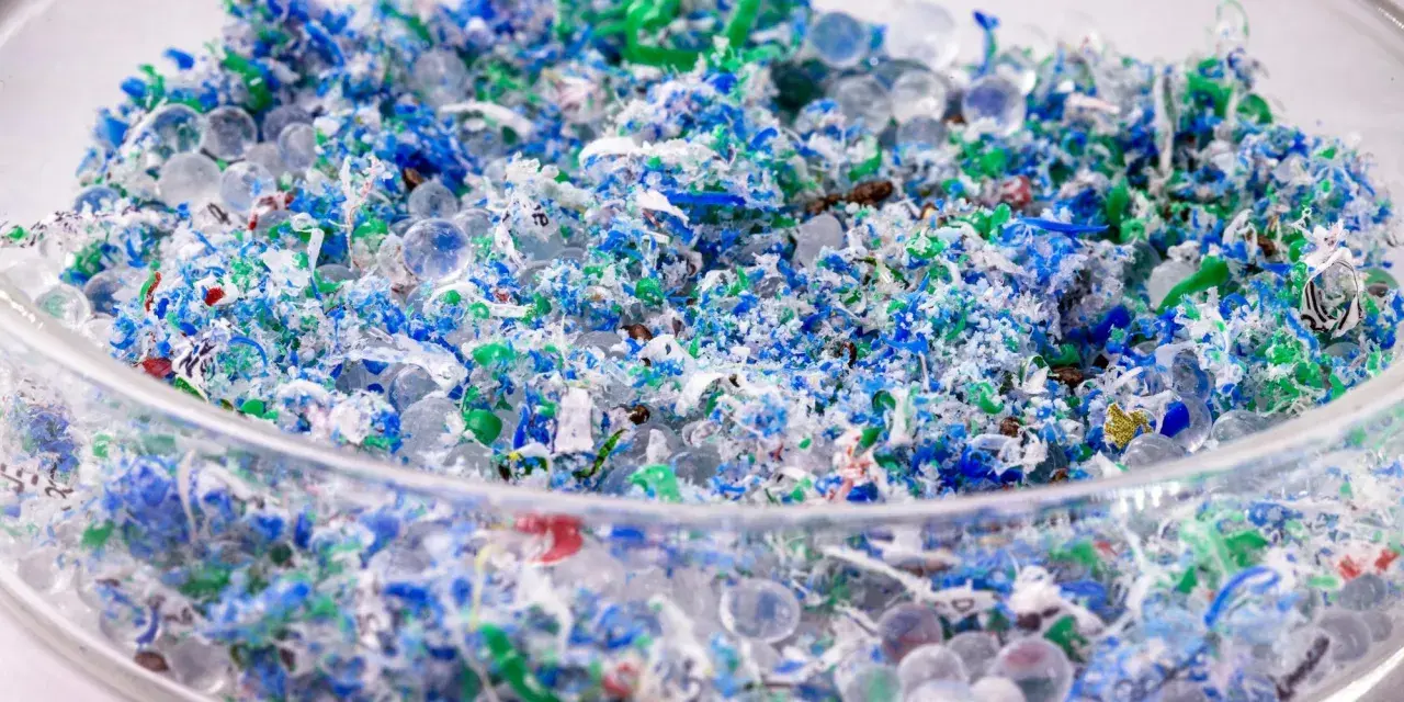 Kolorowe fragmenty plastiku i przezroczyste kuleczki, przypominające mikrokoraliki, tworzą obraz zanieczyszczenia. To mikroplastik w wodzie, widoczny w szklanym naczyniu.