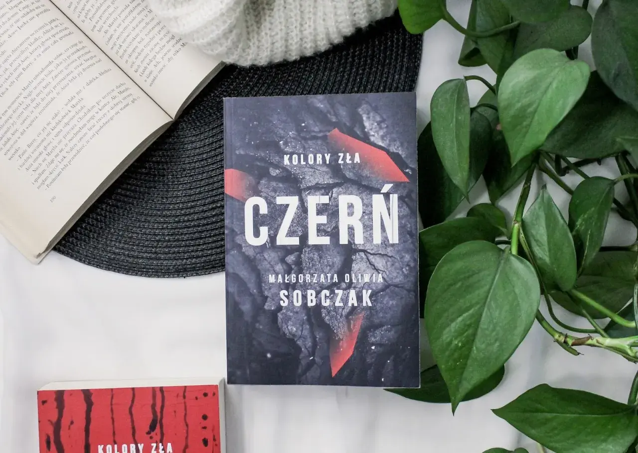 Kolory zła: Czerń. Filmowa okładka książki Małgorzaty Olwii Sobczak, otoczona zielonymi liśćmi i innymi książkami.