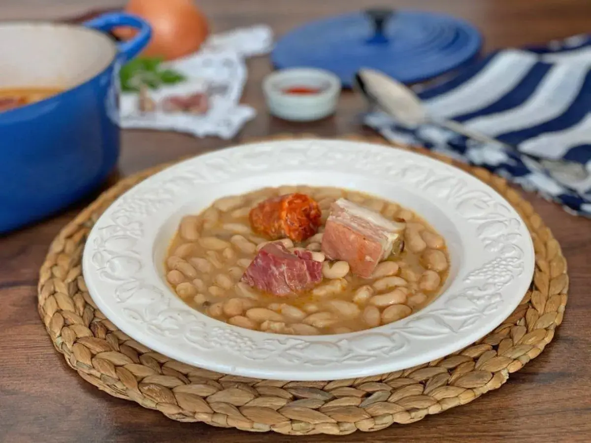 Plato de judías blancas con chorizo y tocino, servido en una vajilla blanca sobre una esterilla de mimbre.