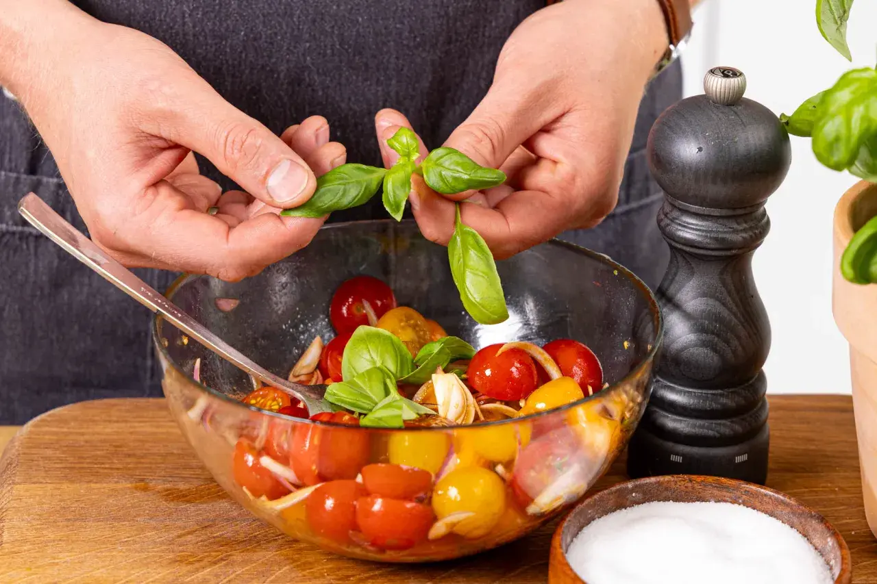 Hände geben Basilikumblätter in eine Schüssel mit Tomaten und Zwiebeln.