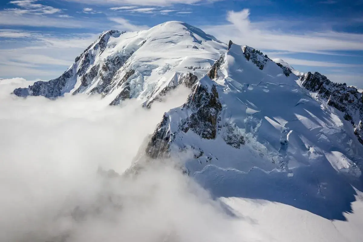 Le Mont Blanc, majestueux et enneigé, émerge des nuages. Des alpinistes escaladent ses pentes glacées sous un ciel bleu éclatant.