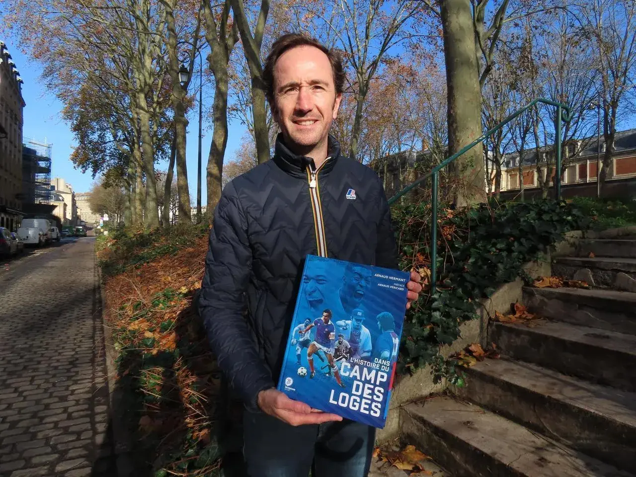 Un homme tient un livre intitulé "Dans l'histoire du Camp des Loges", avec des images de football sur la couverture.