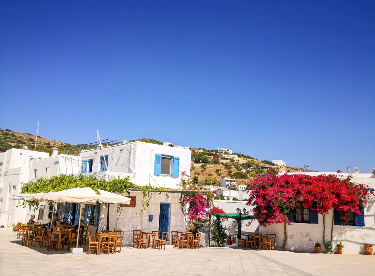 Un angolo di **Grecia con bambini** che giocano felici tra i tavolini di un caffè, sotto bouganville fiorite e il cielo azzurro.