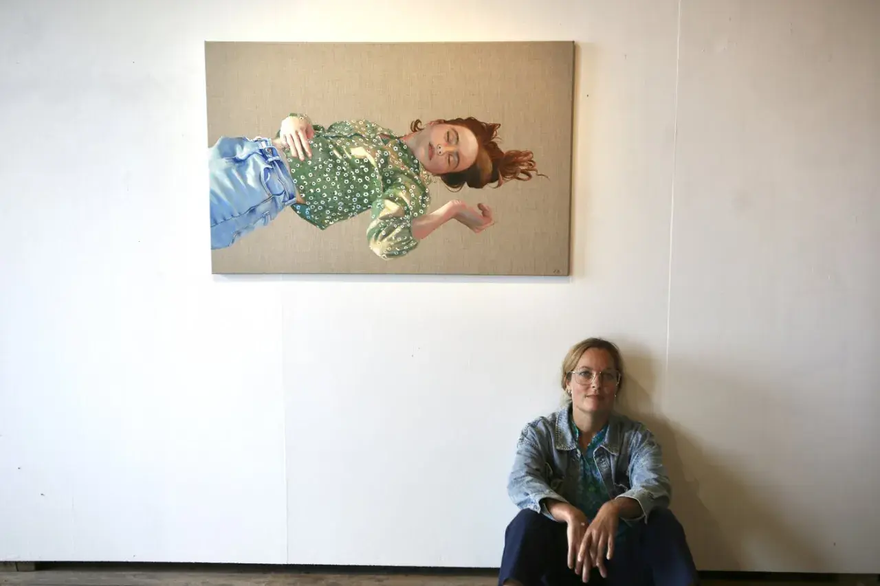 Konstnären Elin Boberg sitter på golvet framför sin målning av en sovande kvinna i grönt mönstrat linne och blå jeans.