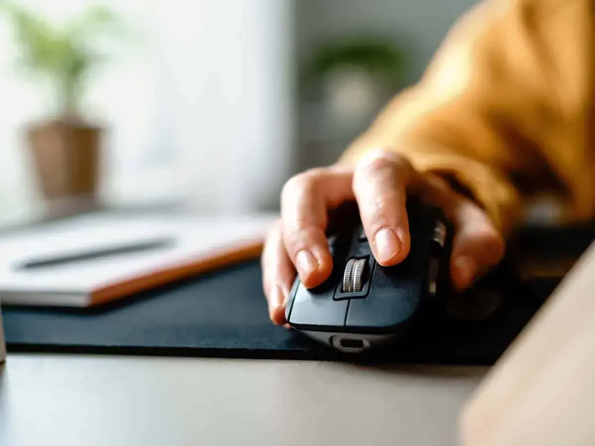 Eine Hand bedient eine Computermaus, die **Maus klickt doppelt**. Im Hintergrund ist eine Pflanze zu sehen.