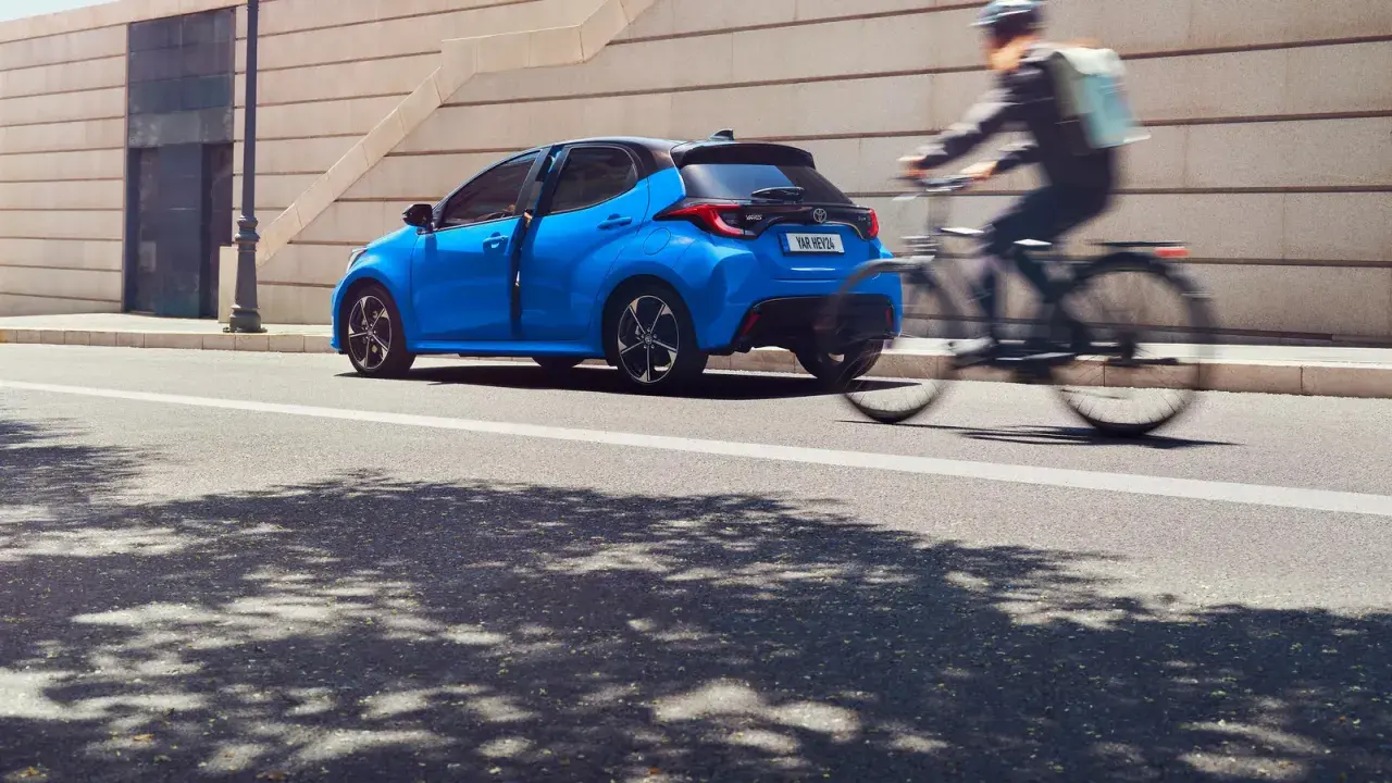 Una Toyota Yaris blu brillante sfreccia in città, mentre un ciclista con zaino passa velocemente. Diversi modelli di Toyota Yaris sono disponibili per ogni stile di vita.