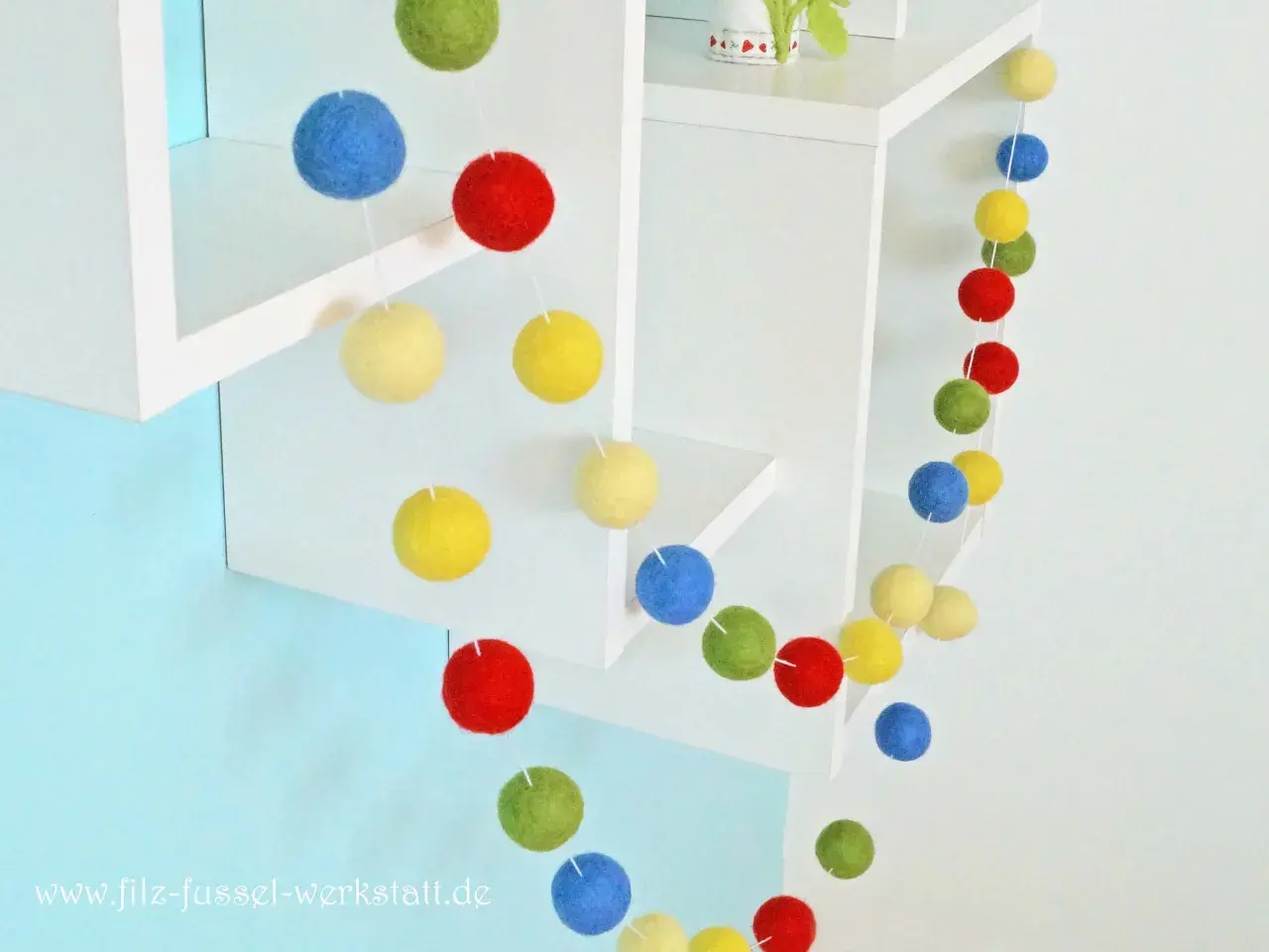 Eine bunte Filzgirlande mit Kugeln in Rot, Blau, Gelb und Grün hängt an weißen Regalen.