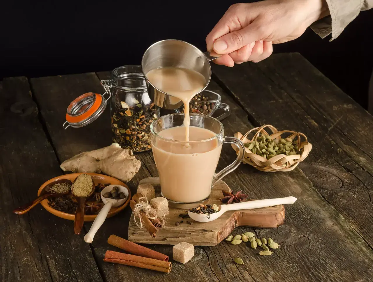 Ręka wlewa mleko do kubka, tworząc aromatyczną herbatę po irlandzku. Wokół przyprawy: cynamon, kardamon, imbir.