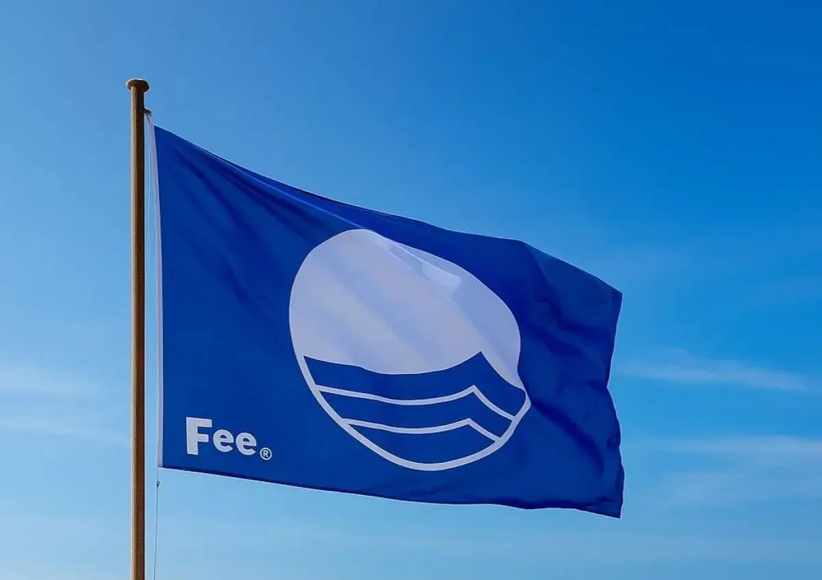 Błękitna flaga z symbolem fal i napisem "Fee" powiewa na maszcie na tle błękitnego nieba.