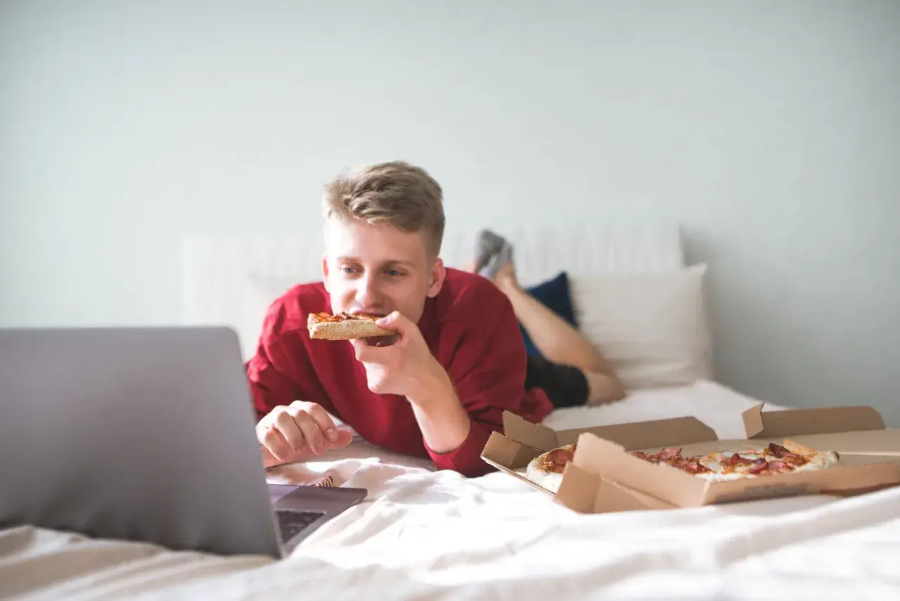 A young man eats pizza in bed while watching a laptop. Is it bad to go to sleep after eating? This scene suggests a late-night snack before bed.