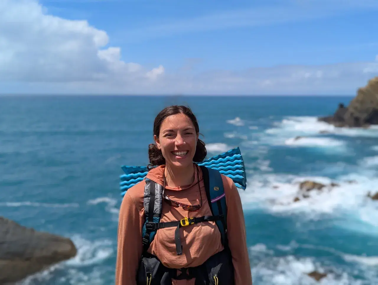 A smiling hiker with a backpack and sleeping pad enjoys the view of the ocean waves crashing on the shore along the Fischerweg trail in Portugal.