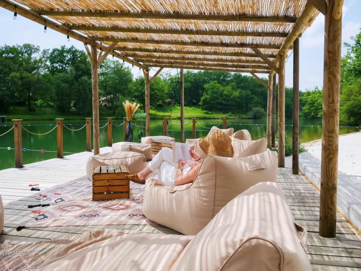 Détente sous le toit de chaume du whaka lodge, au bord d'un lac paisible. Une femme lit, entourée de poufs confortables.