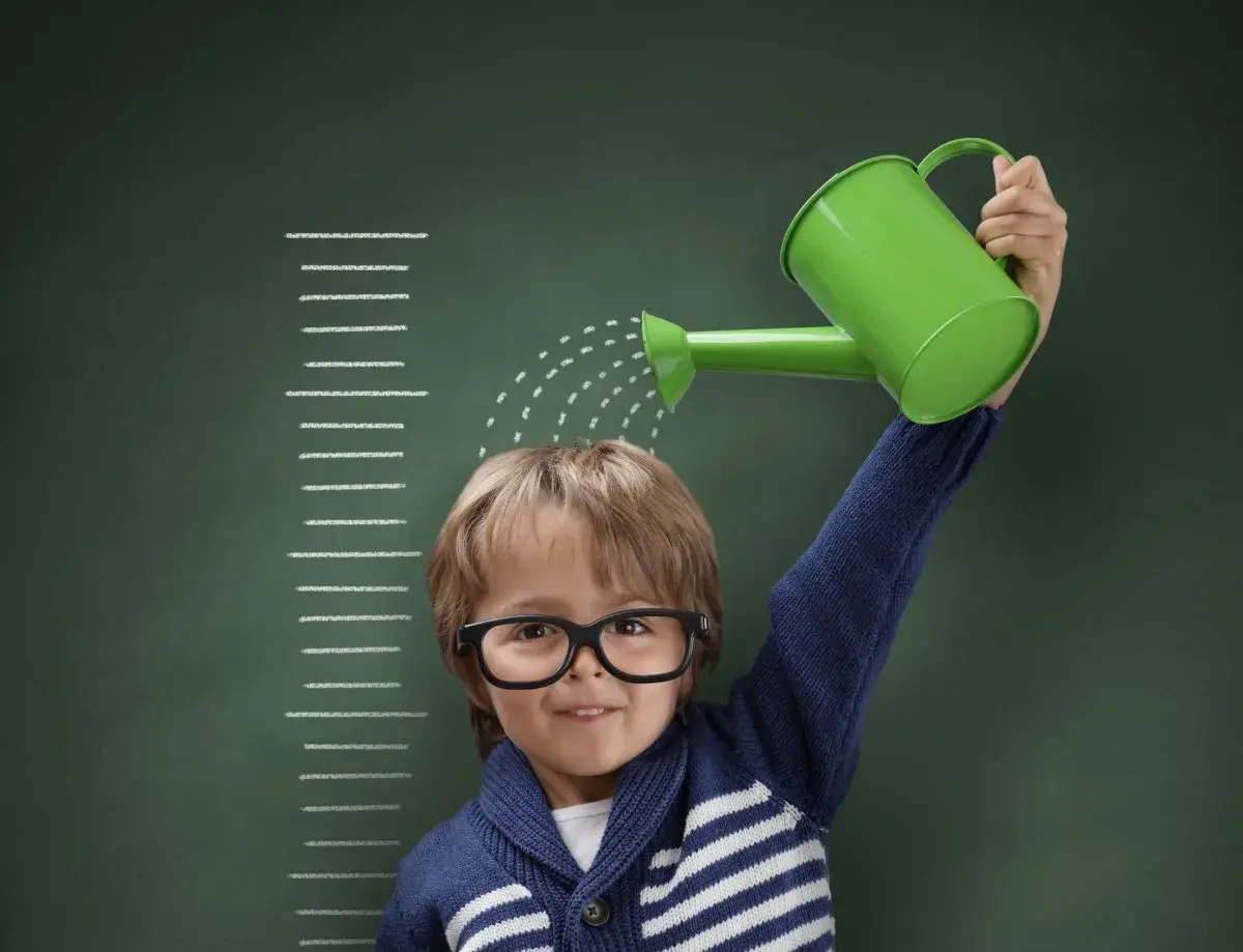 Niño con gafas riega su cabeza con una regadera verde, frente a una tabla de crecimiento que marca el percentil 10.