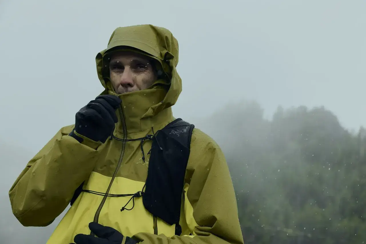 Un homme en veste Gore-Tex jaune et verte, sous la pluie, ajuste son col. Le lavage de cette veste est essentiel pour maintenir son imperméabilité.