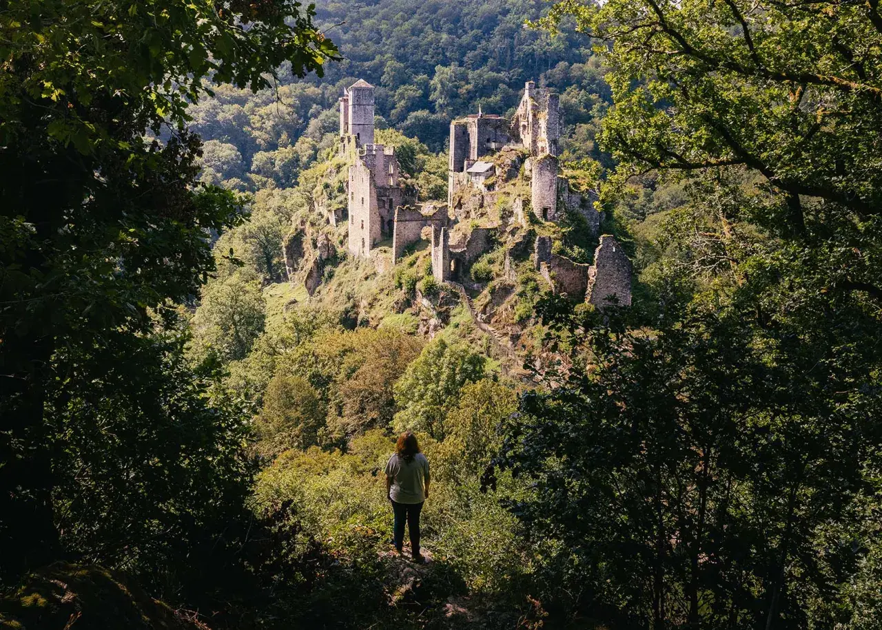 Une personne contemple les ruines d'un château perché sur une colline boisée. Une belle idée de que faire en Corrèze pour les amateurs d'histoire et de nature.