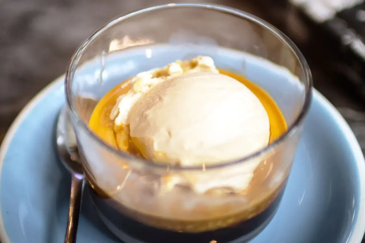 Pyszne affogato: gałka lodów waniliowych w gorącym espresso, podana w szklance na jasnoniebieskim talerzyku.
