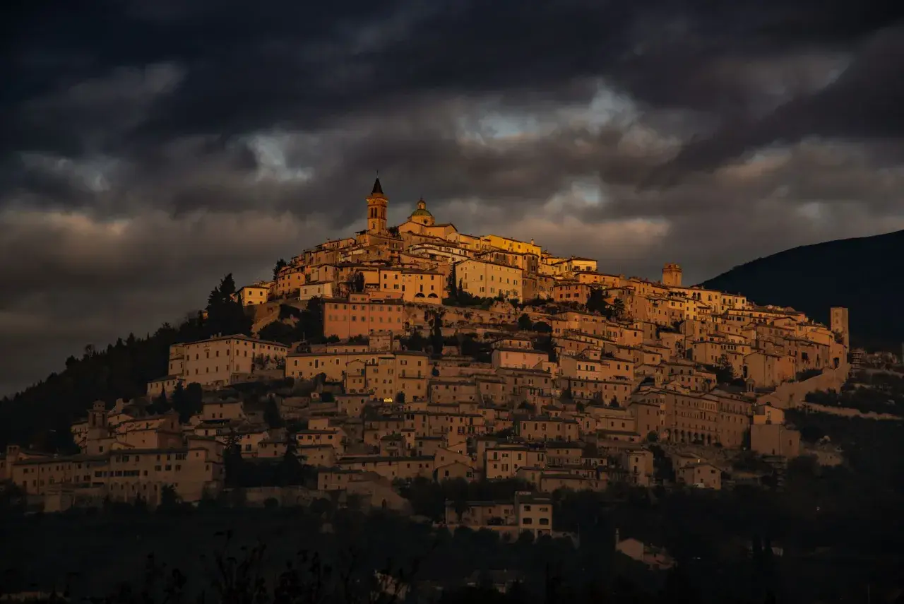 Antico borgo di Trevi, Umbria, illuminato da un sole dorato che filtra tra nuvole scure. Case in pietra si aggrappano alla collina.