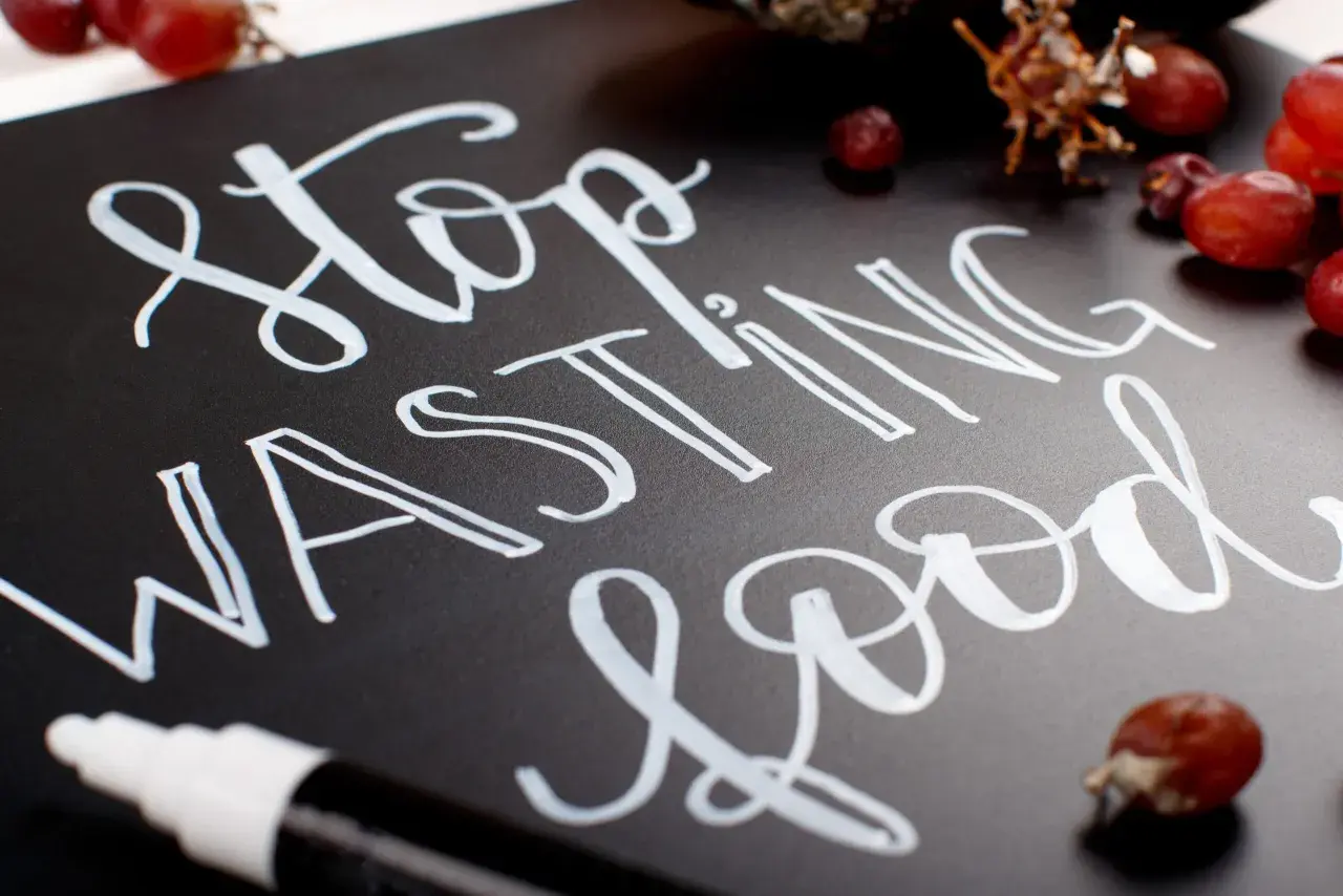 Mensaje "Stop Wasting Food" escrito con diferentes tipos de lettering en una pizarra negra, rodeado de uvas.
