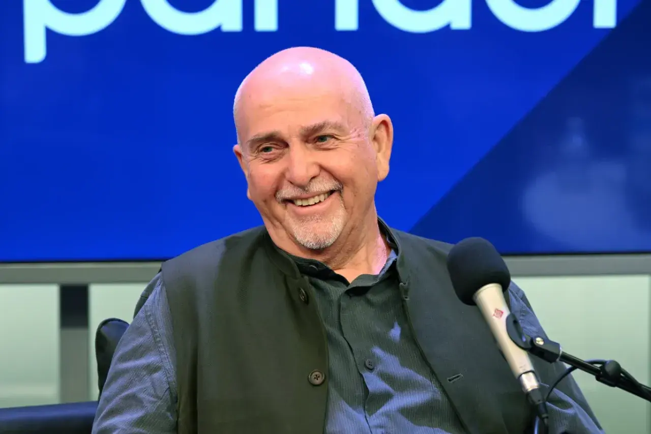 Uśmiechnięty Peter Gabriel, legendarny wokalista, w studiu. W tle niebieskie logo, przypominające o jego bogatej karierze z zespołem Genesis.
