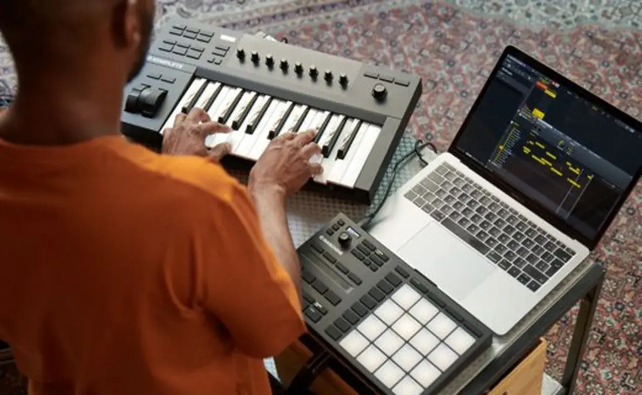 Mann spielt auf einem Keyboard, daneben ein Laptop mit Musiksoftware und ein Pad-Controller. Native Instruments-Geräte für kreative Musikproduktion.