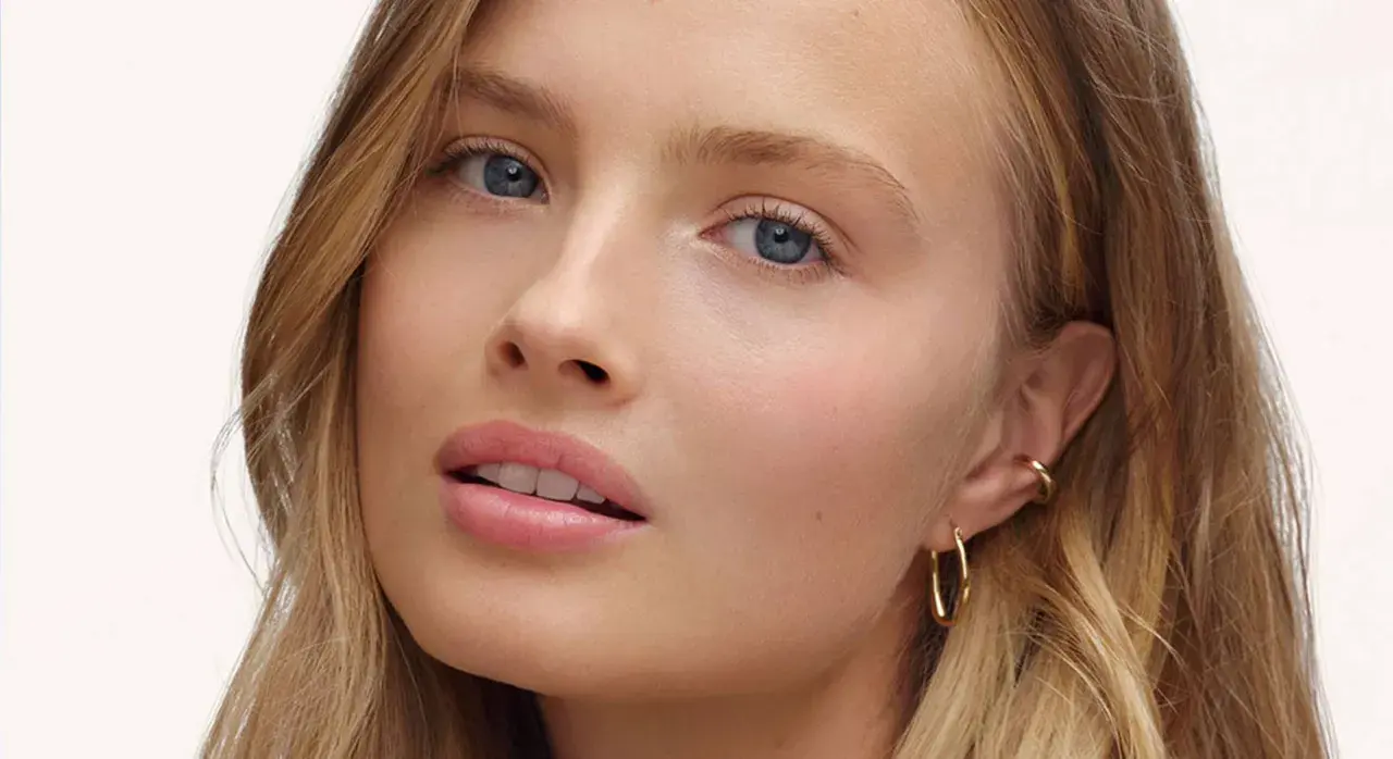 Visage d'une jeune femme aux yeux bleus et aux lèvres rosées, avec un maquillage subtil, style "makeup no makeup". Elle porte des boucles d'oreilles dorées.
