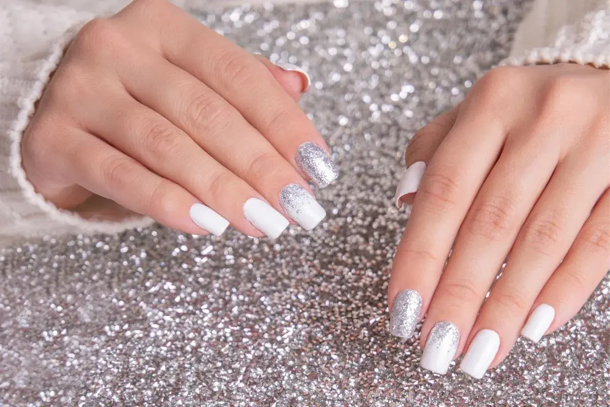 Dłonie z białym french manicure i brokatem na paznokciach, na tle srebrnego brokatu.