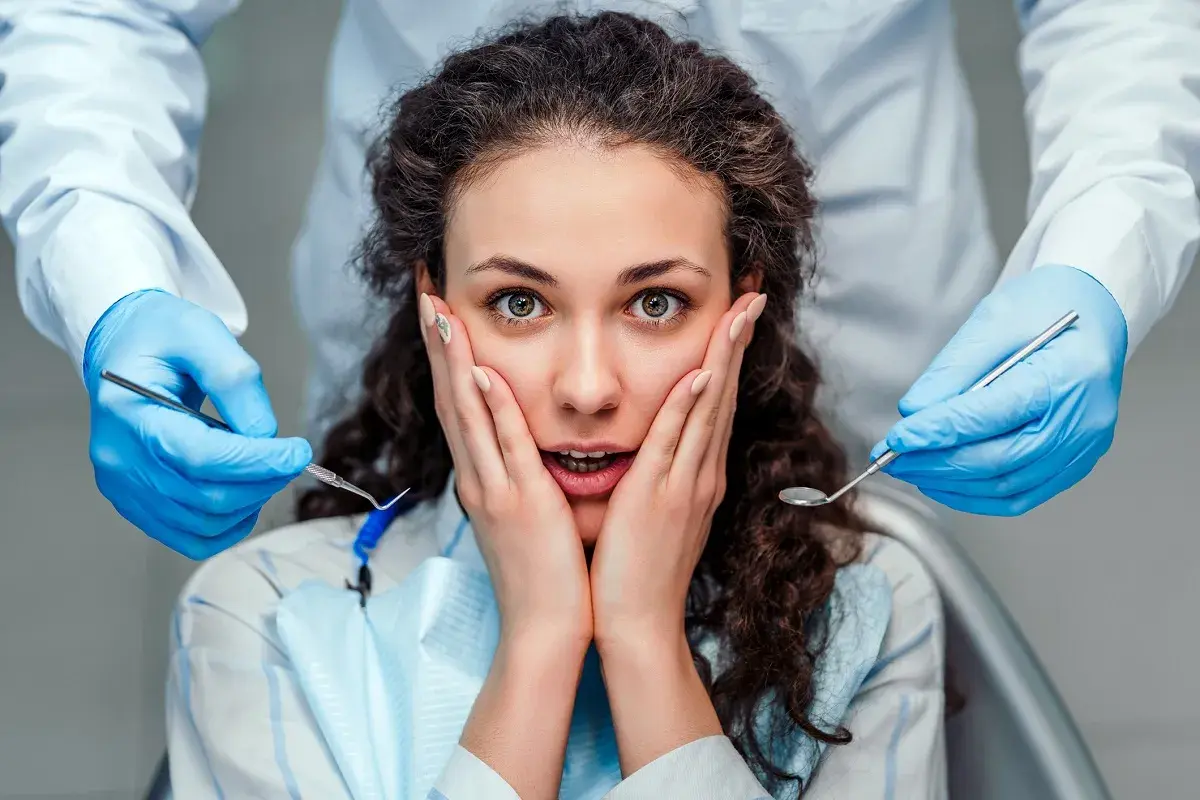 Przerażona kobieta z dłońmi na policzkach, otoczona przez dentystyczne narzędzia. Jej oczy wyrażają strach przed dentystą.