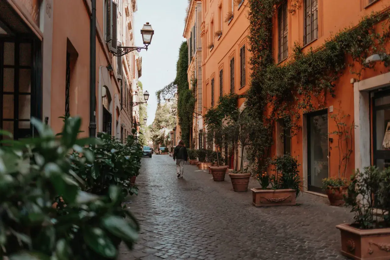 Eine Person geht durch eine schmale Gasse in Rom, gesäumt von orangefarbenen Gebäuden und Pflanzen. Ein perfekter Ort für Rom Geheimtipps.