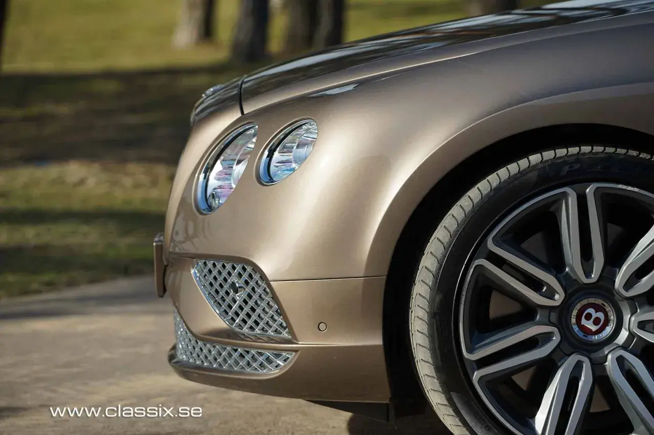 Detaljbild av en lyxig, brun Bentley till salu. Framhjulet och strålkastaren syns tydligt.