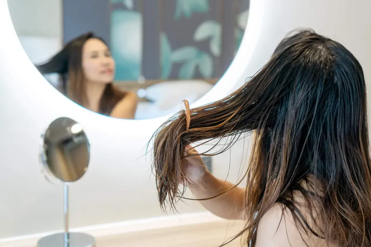 Una joven se peina el pelo mojado frente a un espejo. El pelo adolescentes, largo y castaño, cae en mechones.