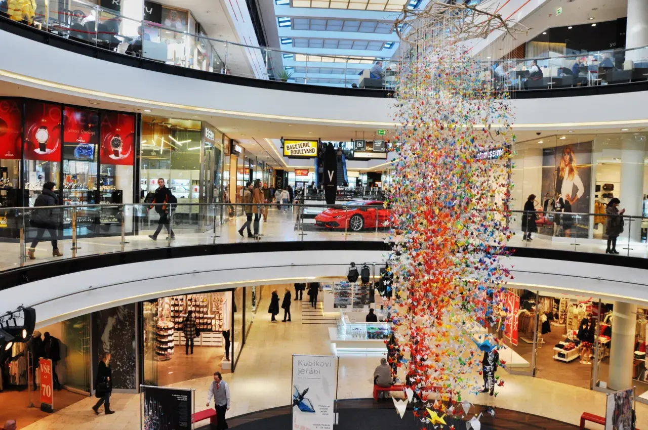 Bunte Papierkraniche schmücken ein Einkaufszentrum in Prag. Menschen bummeln durch die Gänge, vorbei an Geschäften und einer roten Sportwagen-Auslage.