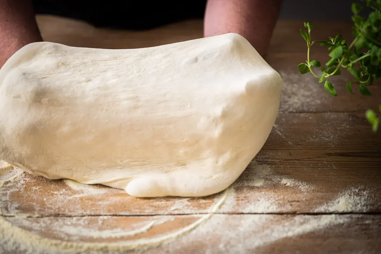Frischer Teig für Pizza Semola wird auf einem Holztisch geknetet, bestäubt mit Mehl. Kräuter liegen daneben.