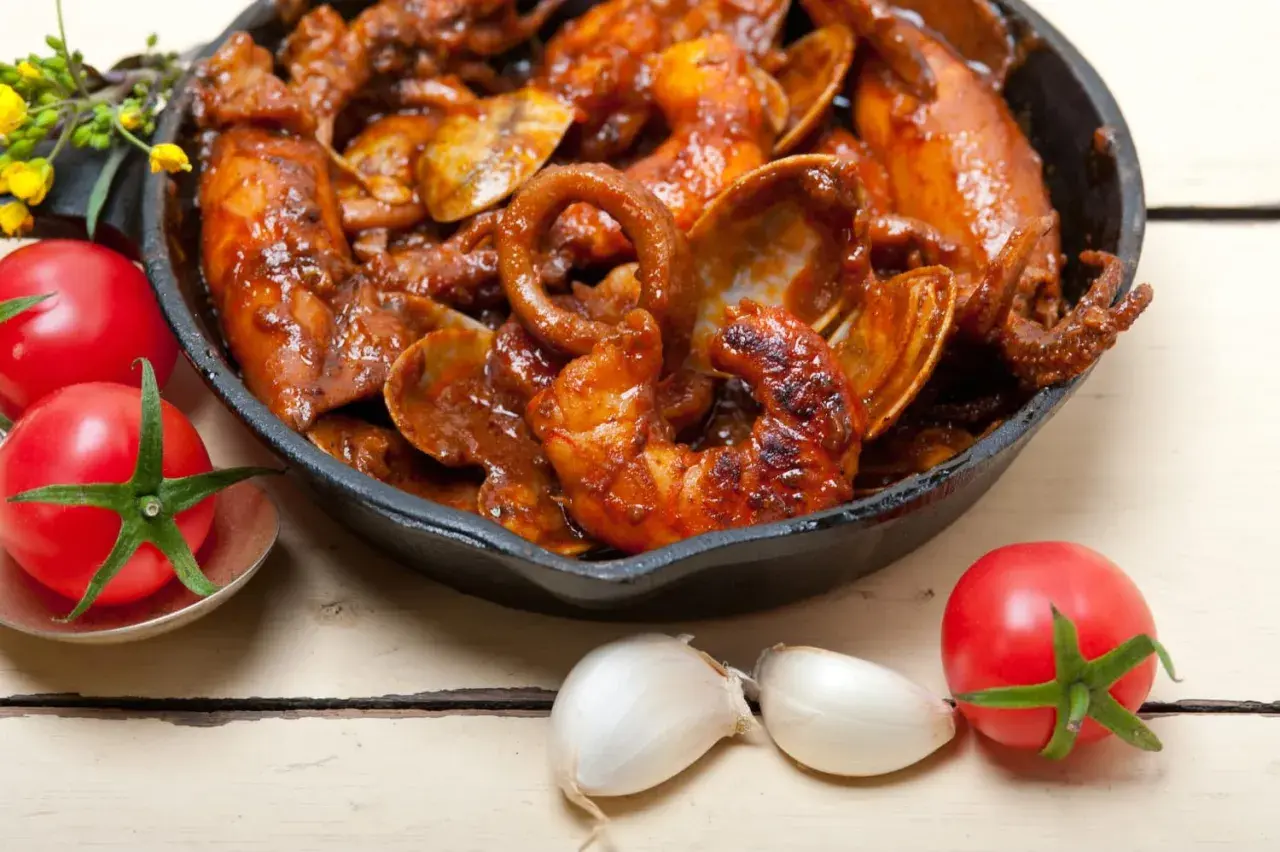 Un plat de calamars à l'armoricaine mijoté dans une sauce rouge, servi dans une poêle en fonte, avec des tomates et de l'ail à proximité.