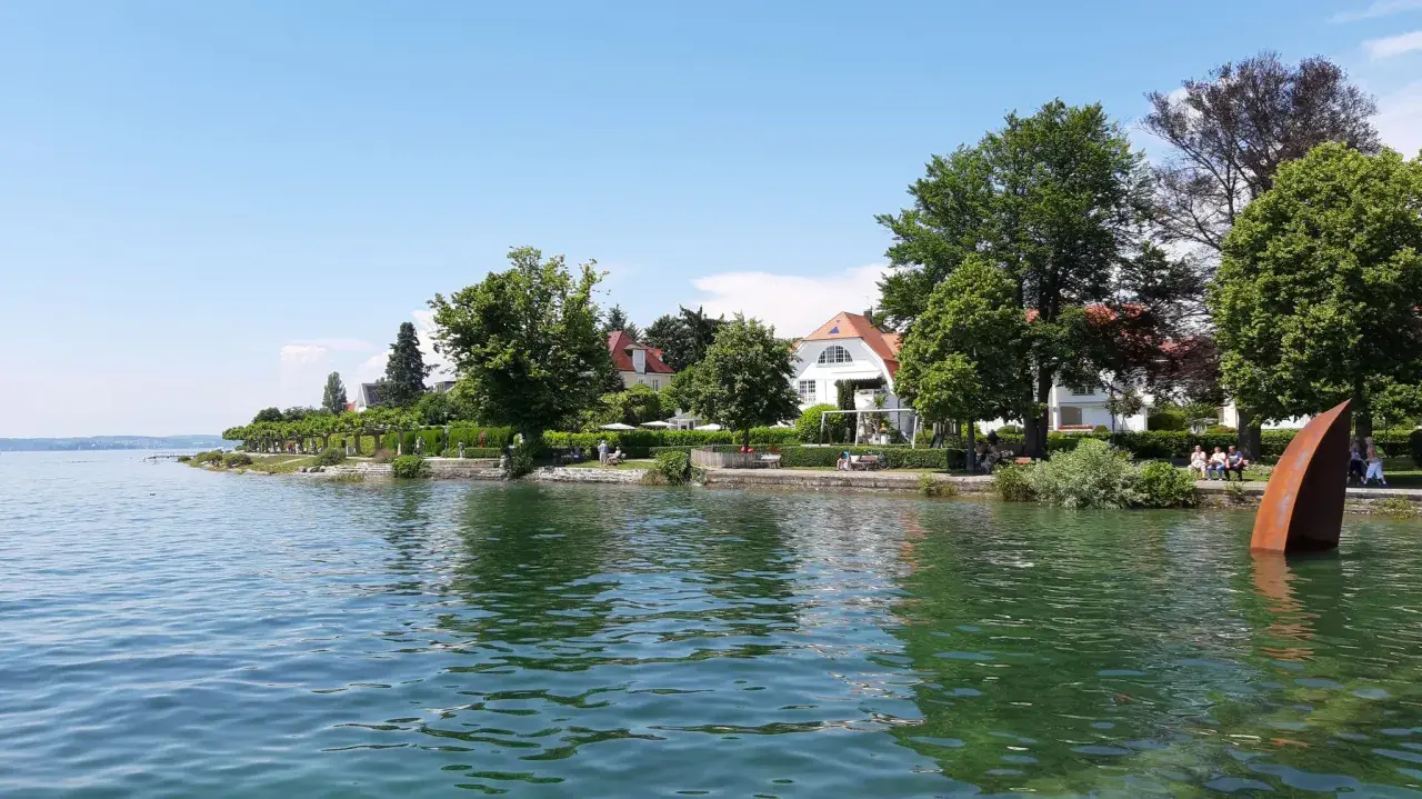 Die Hagnau Promenade am Bodensee mit weißen Häusern, grünen Bäumen und einer rostigen Skulptur im Wasser.
