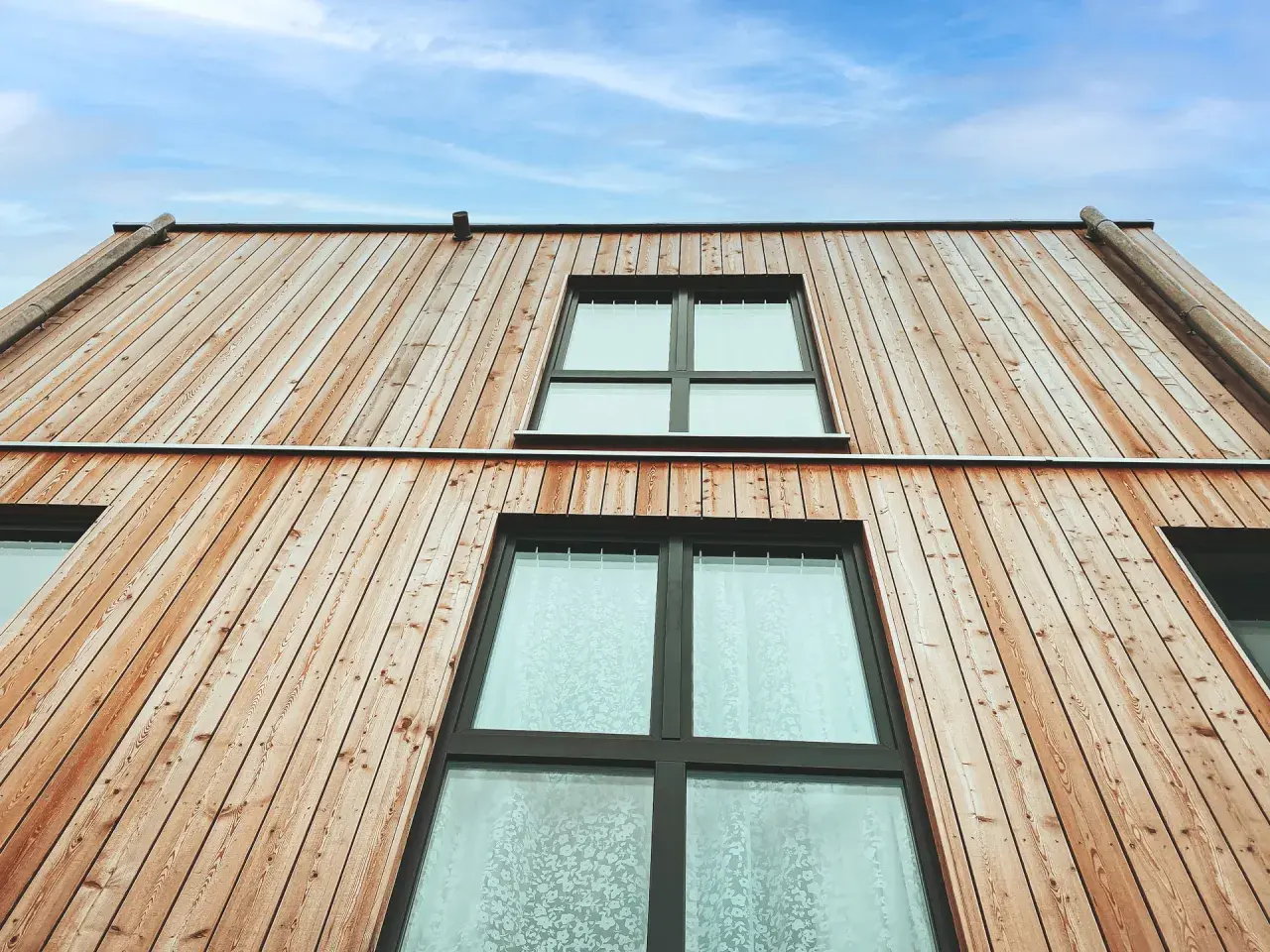 Moderne Holzfassaden Beispiele: Natürliche Holzverkleidung mit großen Fenstern unter blauem Himmel.