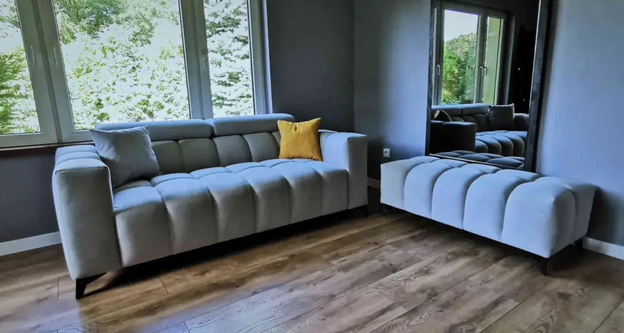 Nowoczesna szara sofa i pufa, ozdobione żółtą poduszką. To meble od najlepszych producentów mebli tapicerowanych, idealne do stylowego salonu.