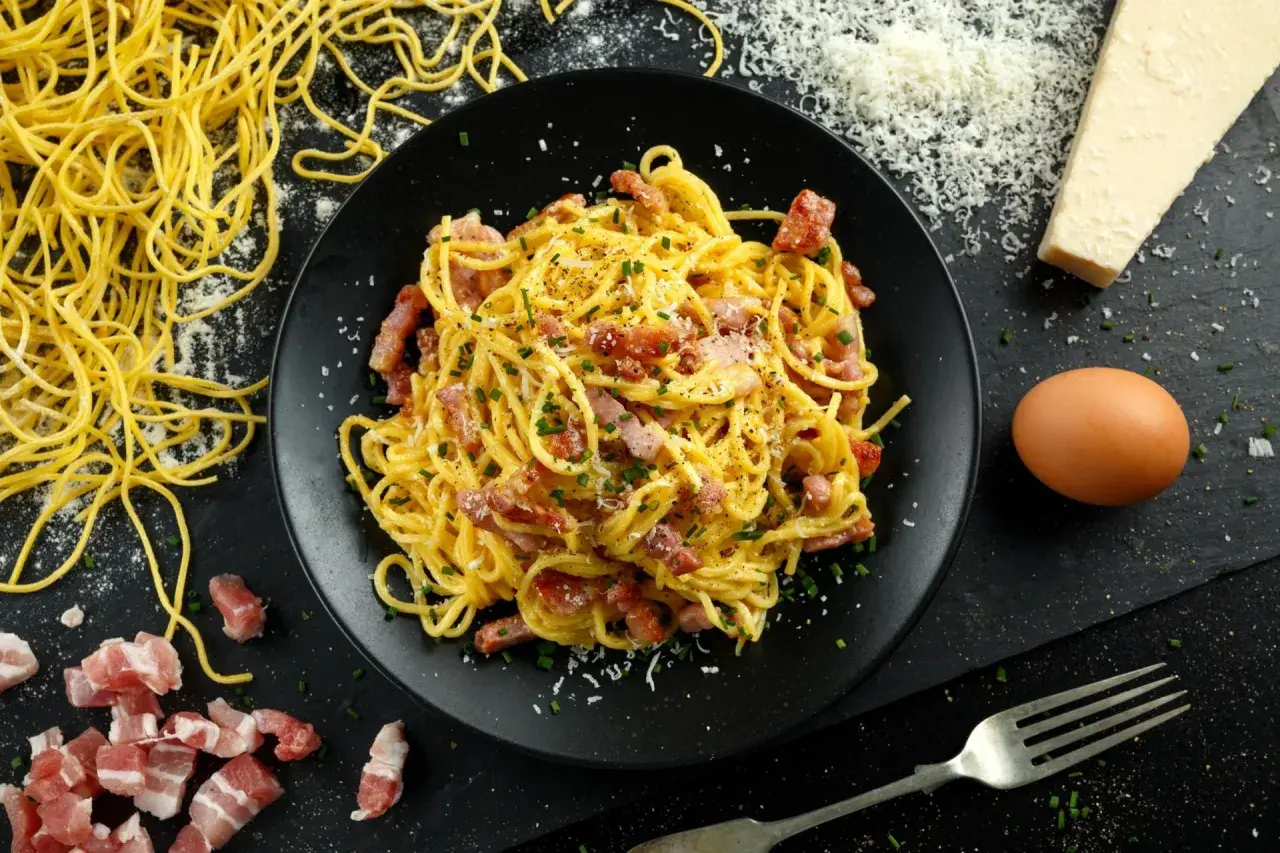 Pyszne spaghetti carbonara na talerzu, obok surowy boczek, jajko i ser. Zastanawiasz się, ile kalorii ma spaghetti carbonara?