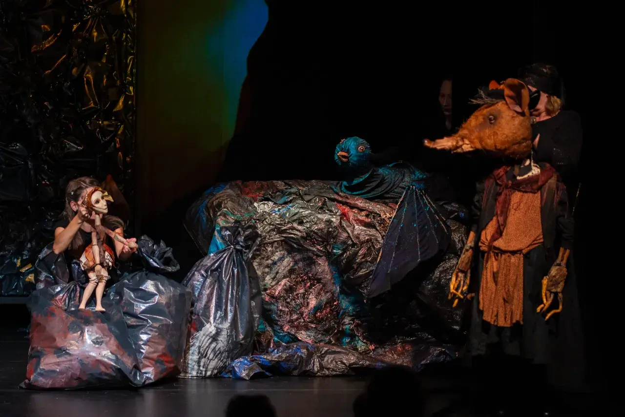 Na scenie teatru lalki i aktora w Łomży, dziewczynka animuje drewnianą lalkę, obok niej ptak i postać z głową zwierzęcia.