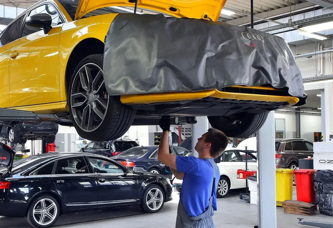 Mechanik pracuje pod żółtym Audi, sprawdzając, ile kosztuje wymiana oleju silnikowego w samochodzie. W tle inne auta.