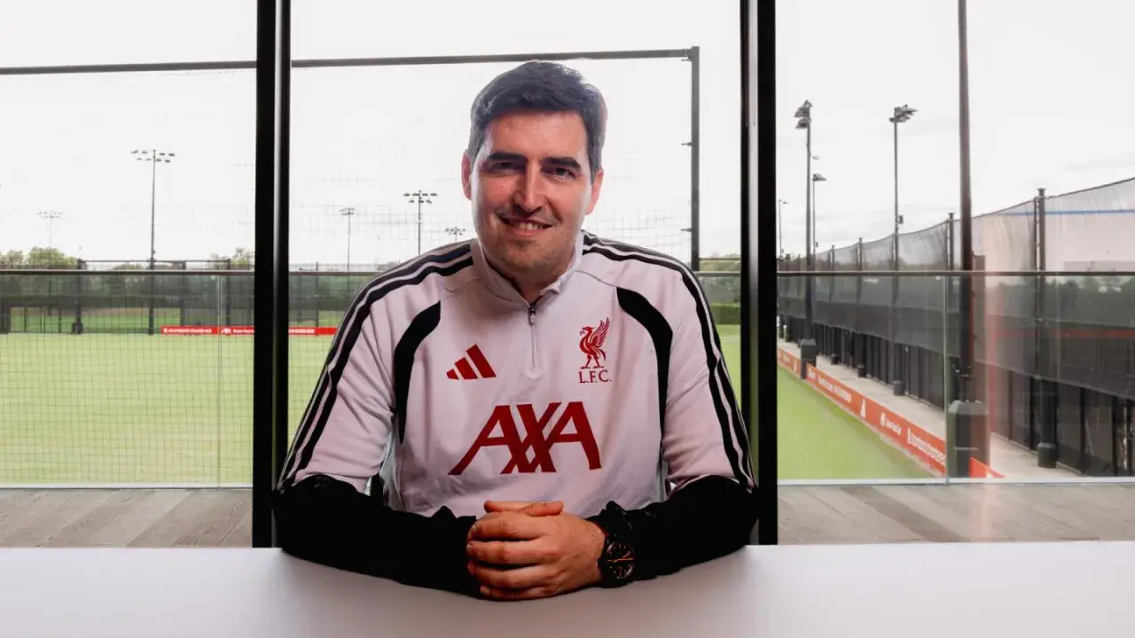Uśmiechnięty trener Liverpoolu w stroju treningowym z herbem klubu i logo AXA. Za nim boisko treningowe.