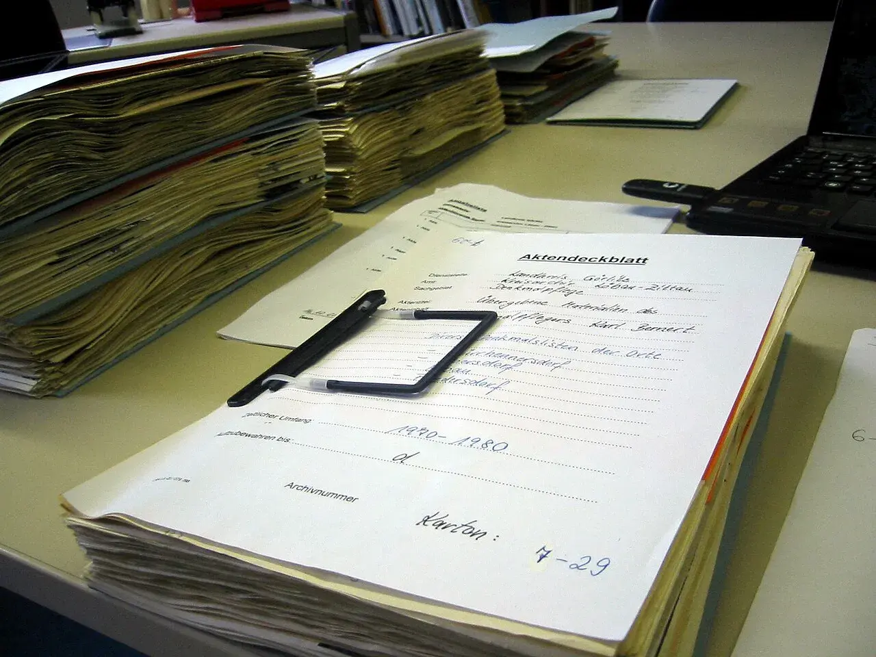 Stosy dokumentów, długopis i laptop na biurku. Na jednym z dokumentów widnieje napis "Akten-dossier co to", sugerujący porządkowanie akt.
