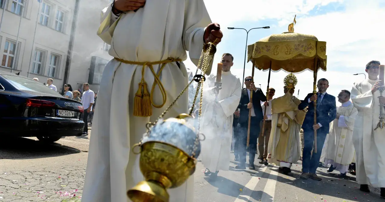 Procesja Bożego Ciała z kadzidłem i monstrancją. Uczestnicy w białych szatach idą ulicą.