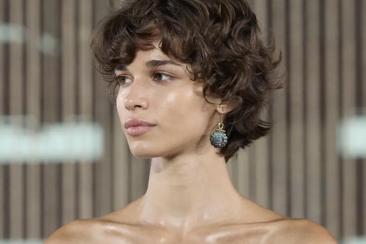 Mujer con pelo corto fino y escaso, rizado y castaño. Luce un pendiente colgante con una piedra grisácea.