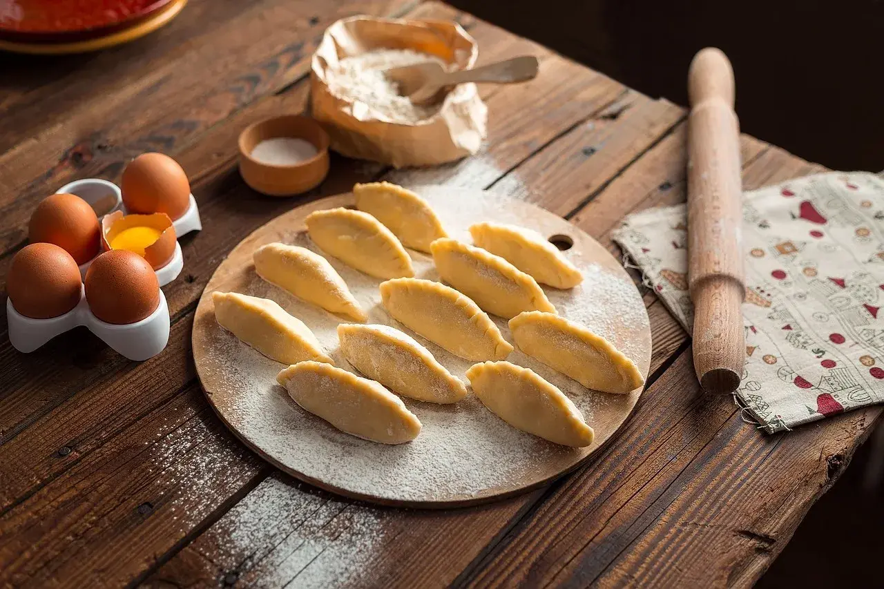 Surowe pierogi ułożone na desce, obok jajka, mąka i wałek.