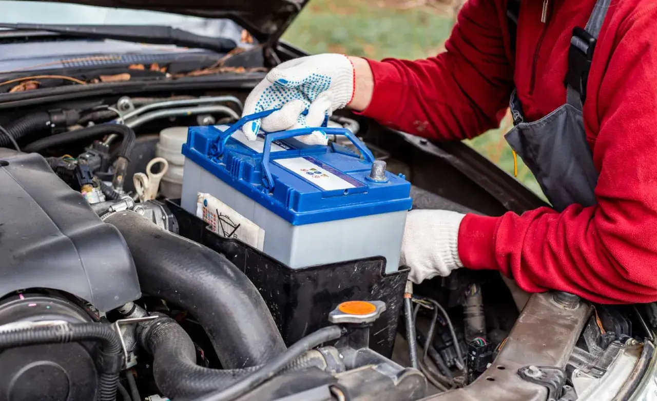 Persona con guantes blancos manipula una batería de coche azul y gris, lista para cargar batería coche.