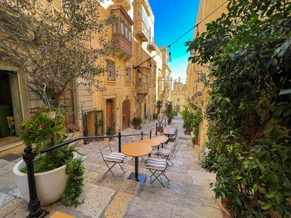 Malta: co warto wiedzieć? Urokliwa uliczka z kawiarnianymi stolikami i krzesłami, otoczona kamiennymi budynkami i zielenią.