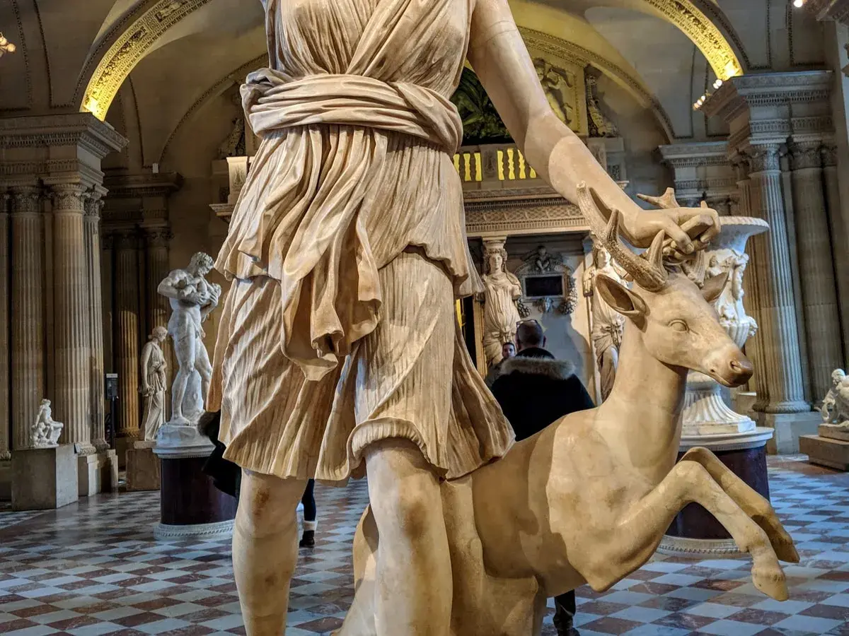 Statue de Diane chasseresse, qui est Artémis, tenant un cerf. L'œuvre est dans un musée, avec d'autres sculptures en arrière-plan.